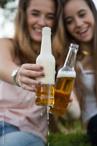 Two young laughing women on lawn in park toasting with beer bottles having fun. 