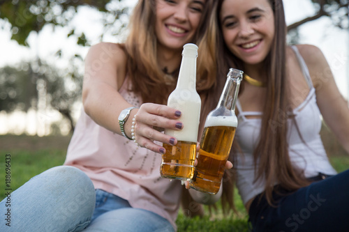 Two young laughing women posing on lawn in park and toasting with beer bottles having fun. 