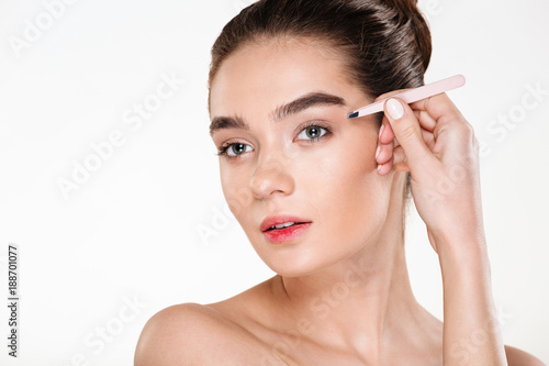 Close up studio portrait of young brunette woman with soft skin plucking eyebrows with tweezers isolated over white background
