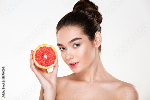 Portrait of pretty half-naked woman with natural makeup holding red orange near her face and looking on camera over white background