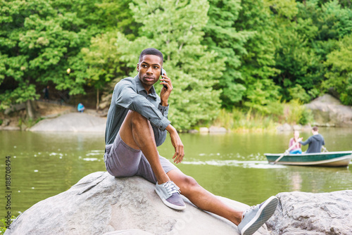 Young African American Man wearing gray long sleeve shirt, shorts, sneakers, sitting on rocks by lake at Central Park in New York, relaxing, talking on cell phone. People rowing boats on background..