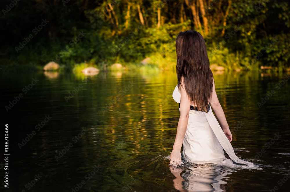 Fototapeta premium A long-haired fit girl with white dress walking through the river touching water