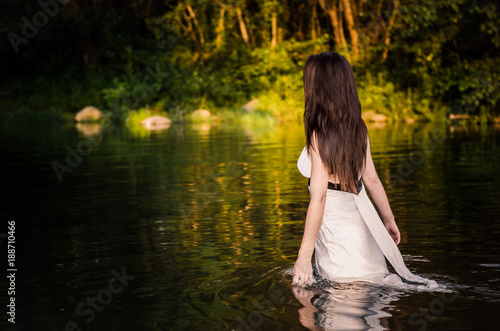 Fototapeta Naklejka Na Ścianę i Meble -  A long-haired fit girl with white dress walking through the river touching water