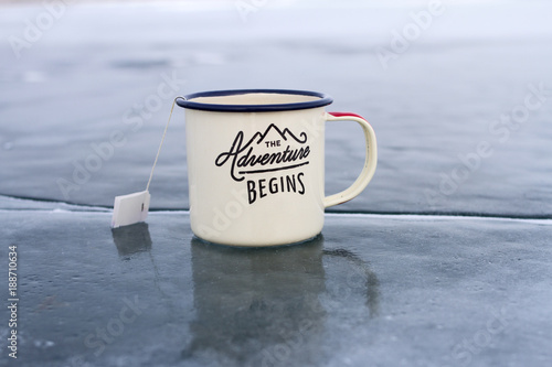 Enameled mug with hot tea and a label with an empty space for an inscription from a tea bag on the blue clear ice of Lake Baikal in winter on a sunny day.