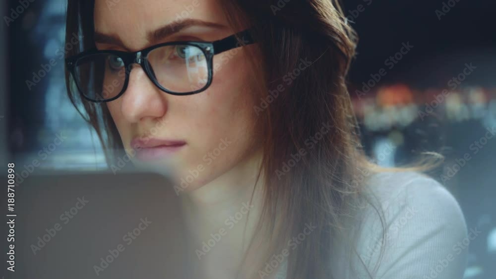 Beautiful young businesswoman wearing glasses and working at night using modern computer touchscreen, confident female employer working at new office with panoramic view of night city