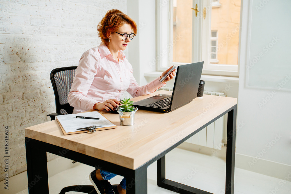 Red hair mature business woman in glasses sitting on wooden table with plant, disposable cup of coffee and paper, holding smartphone and working with laptop near window. Red hair mature business woman in glasses sitting on wooden table with plant, disposable cup of coffee and paper, holding smartphone and working with laptop near window.