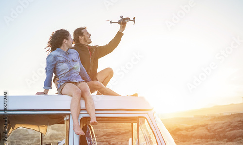 Happy couple using a drone sitting on top of vintage minivan at sunset
