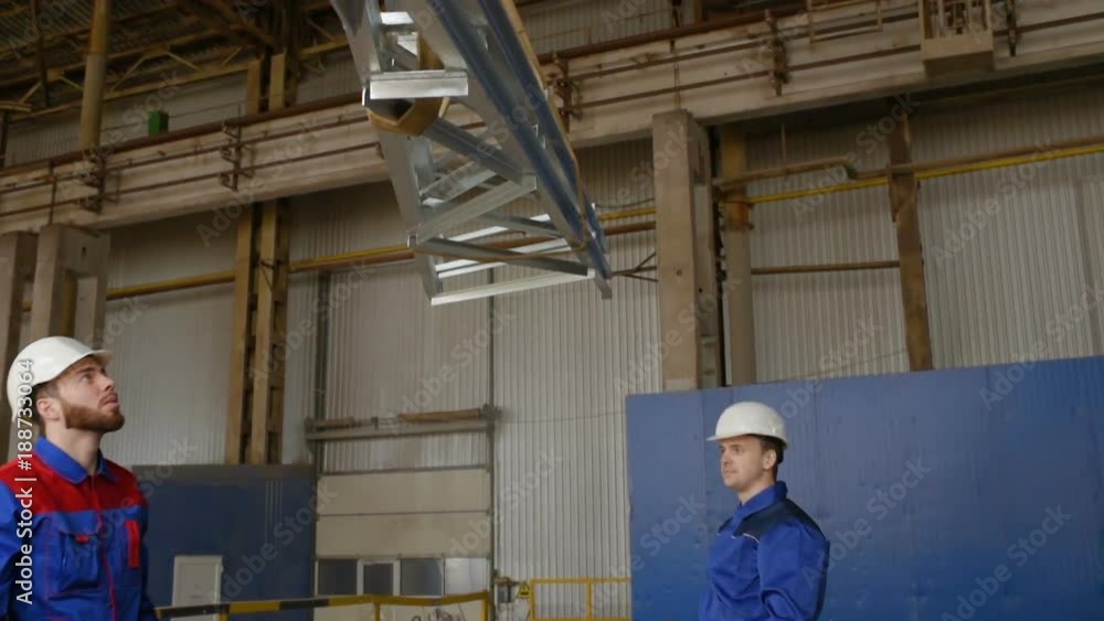 Two workers in white construction helmets load industrial products on the hook of the crane against the background of production Two workers in white construction helmets load industrial products on the hook of the crane against the background of production