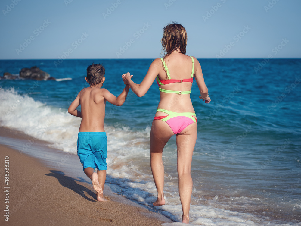 Mom and son are running along the sea shore. They are holding hands and moving away from the camera.