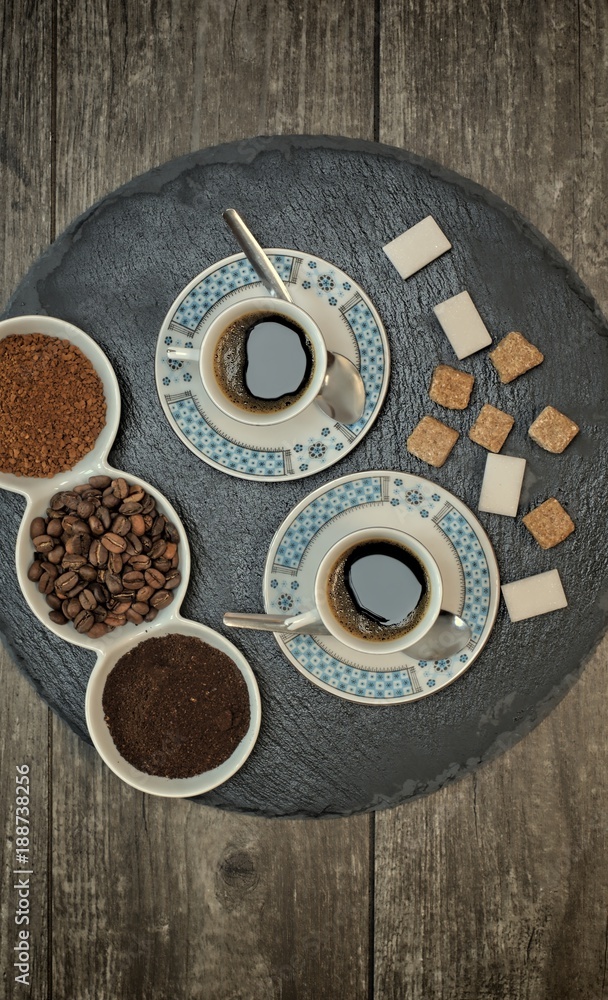Two cups of coffee, coffee beans, ground coffee and instant coffee on a dark slate stand and wooden background.