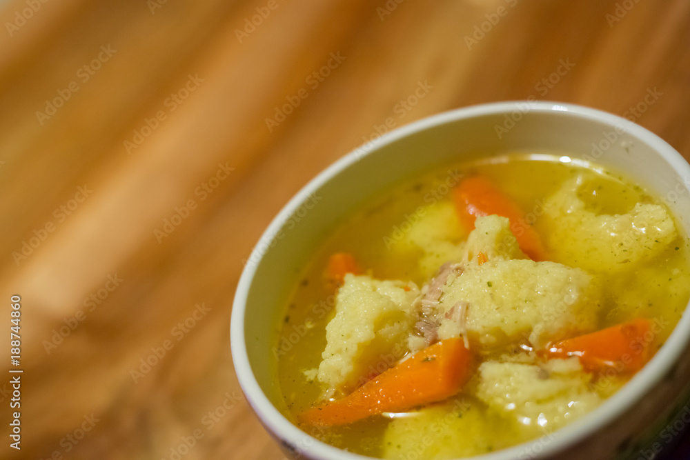 Hot chicken soup with dumplings and carrot in a small porcelain bowl on wooden table Hot chicken soup with dumplings and carrot in a small porcelain bowl on wooden table
