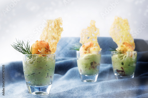 avocado cream guacamole with tiger prawn shrimps and crisp cheese cracker in three glasses, festive appetizer or party snack on a blue tablecloth background with blurry bokeh lights, copy space