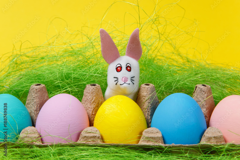 Row of five colorful pastel monophonic painted Easter eggs in cardboard tray, white bunny in green grass on yellow background. Happy Easter concept. Copy space for advertisement. With place for text.