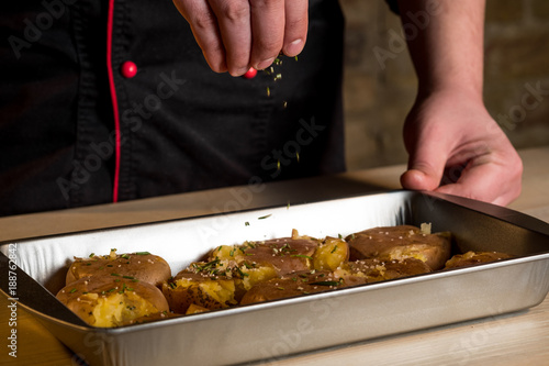 Chef hands cooking potatoes in baking tray with herbs, salt, oil and spices. Step homemade recipe. Food dark wooden background.