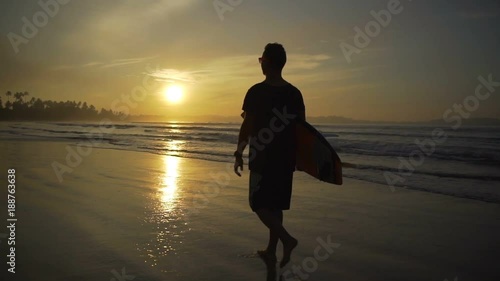 Young man walking on sandy beach holding longboard sunset rapid slow motion