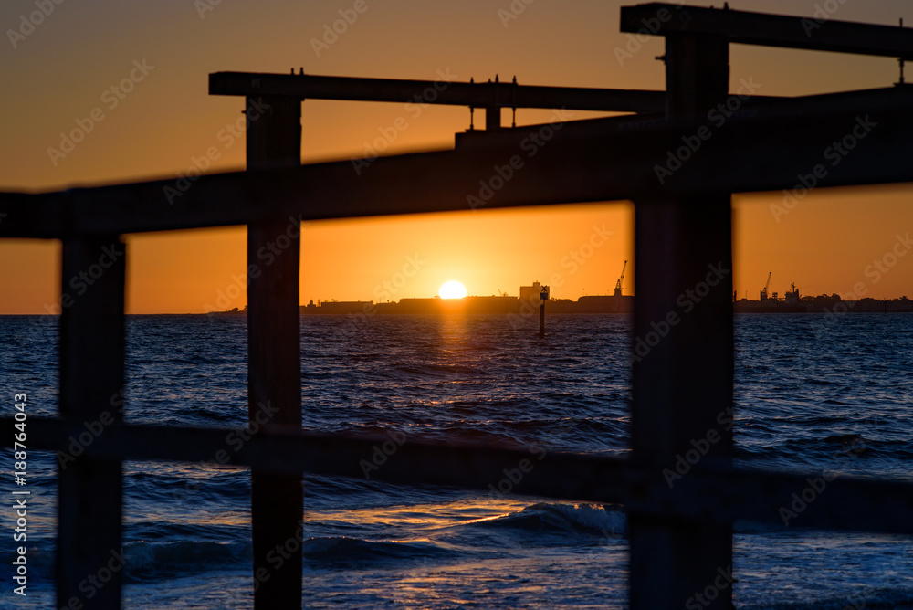 The sun sets over the horizon of Port Phillip Bay with a silhouetted wooden boat launch structure in the foreground. Melbourne Australia