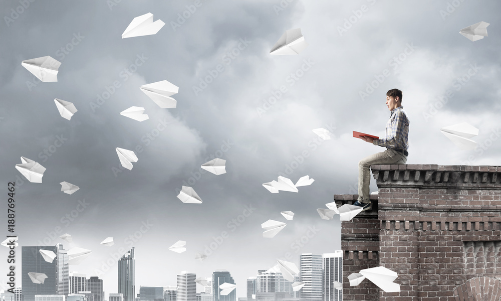 Handsome student guy on roof edge reading book and paper planes fly around Handsome student guy on roof edge reading book and paper planes fly around
