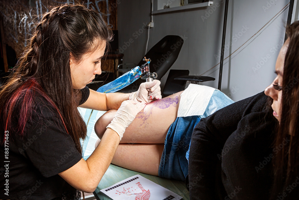 Young woman tattoo artist doing tattoo with bird tattoo machine on  dark-haired beautiful woman in dark workshop Stock Photo | Adobe Stock