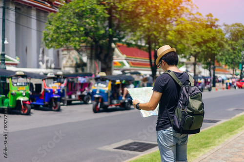 Young asian man traveler with sky blue backpack and hat looking map find the way with tuk tuk thailand background from wat pho at Bangkok Thailand