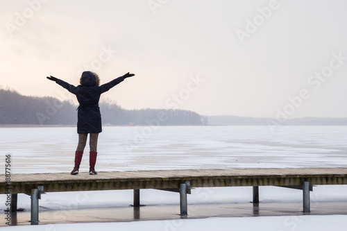 Young happy woman standing alone on footbridge at the lake ice and enjoying freedom. Girl back. Sunrise in cold atmosphere in the morning. Nature after snow blizzard. Waiting for a new day in winter.