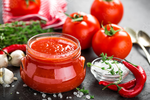 Tomato paste, puree in glass jar and fresh tomatos on dark background. Hot vegetable sauce with chili pepper and tomatoes