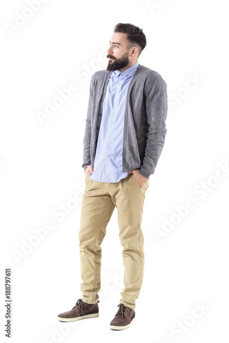 Bearded male fashion model in gray cardigan with hands in pockets looking away. Full body length portrait isolated on white studio background. 