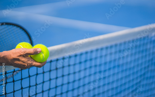 old asian man hold two tennis balls in right hand, selective focus, blurred racket, net and blue tennis court as background, “aging population” concept © angyim