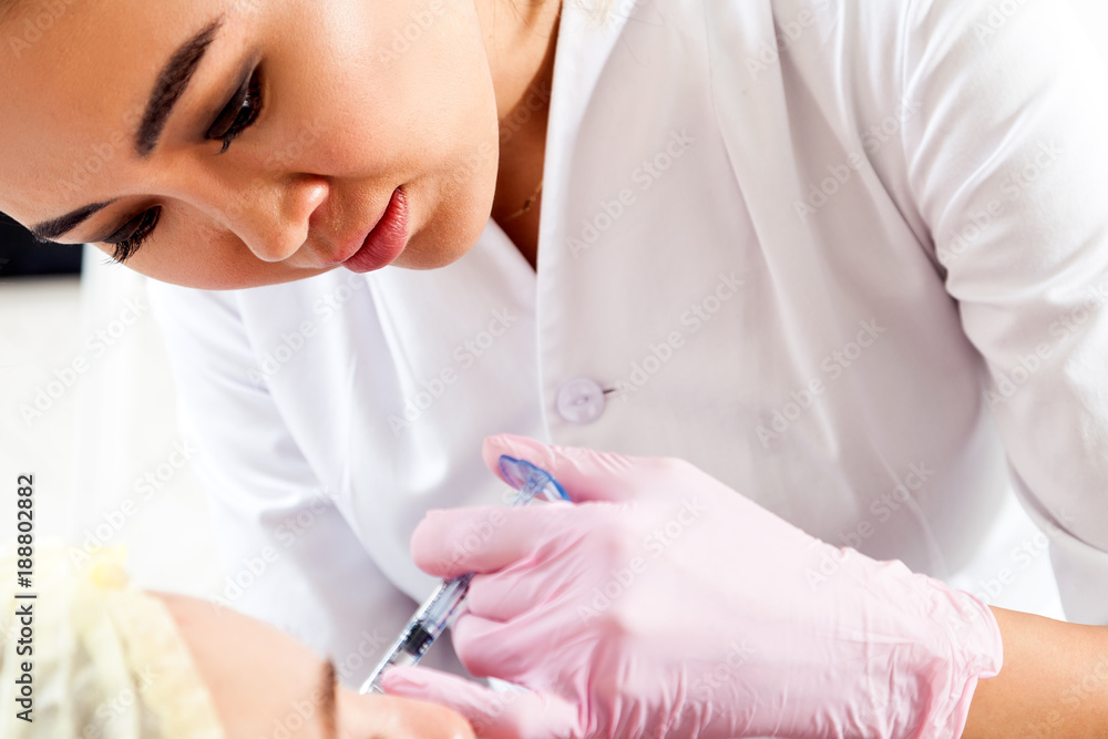 Close-up Young woman doctor cosmetician in white lab coat and sterile gloves injects Botox with syringe into cheekbones Close-up Young woman doctor cosmetician in white lab coat and sterile gloves injects Botox with syringe into cheekbones