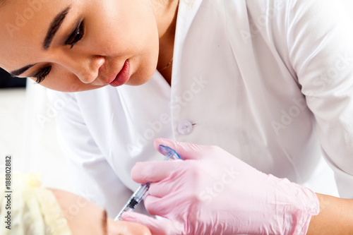 Close-up Young woman doctor cosmetician in white lab coat and sterile gloves injects Botox with syringe into cheekbones
