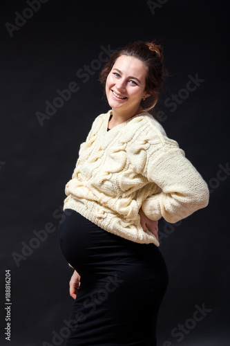 Close-up of a young pregnant woman woman in a knitted milk-colored sweater and a black dress smiling and holding herself behind her back on a black isolated background