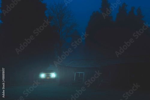 Off road car with headlights on in misty forest near old barn at dusk.