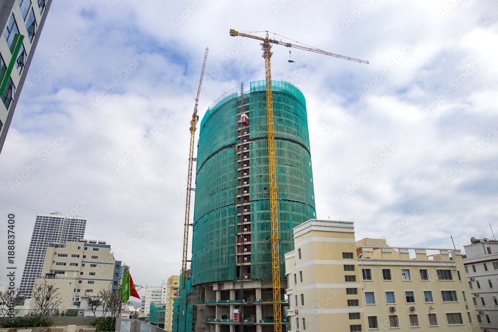 Fototapeta premium construction of a high-rise building in scaffolding and two yellow construction cranes on the background of the blue cloudy sky