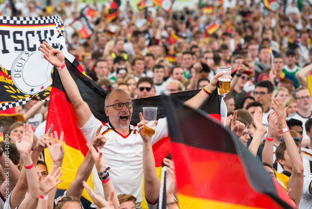 Obraz premium Jubelnder Fußball-Fan mit deutscher Fahne und einem Becher Bier in einer Menge jubelnder Fans beim Public Viewing