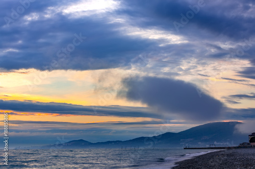 High clouds and fog on the coast, the sun is hidden behind the clouds, on the shoreline are visible the houses of the city on the hills. Beautiful colorful sunset on the Black Sea, Sochi, Russia.