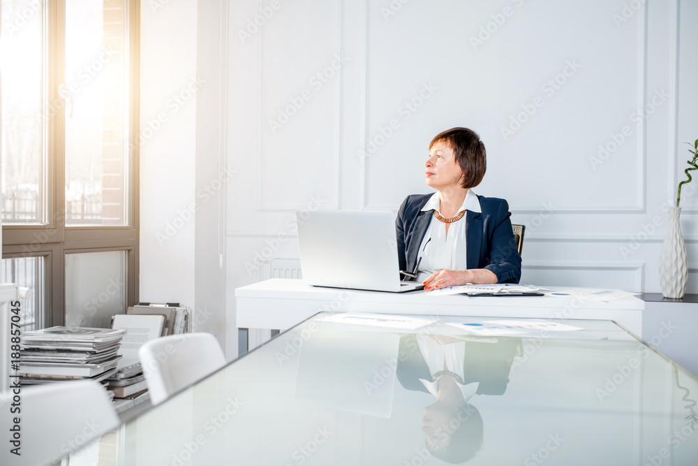 Elegant senior businesswoman dressed in the suit working with laptop and documents at the white office. Wide angle view with copy space