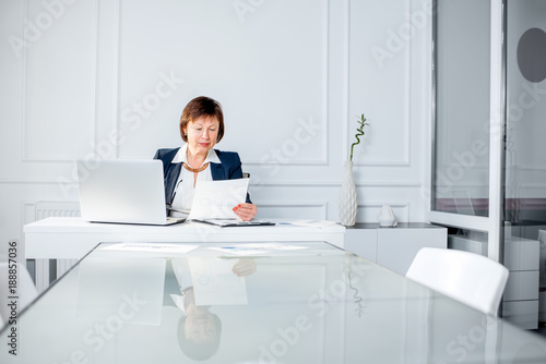 Elegant senior businesswoman dressed in the suit working with laptop and documents at the white office. Wide angle view with copy space © rh2010