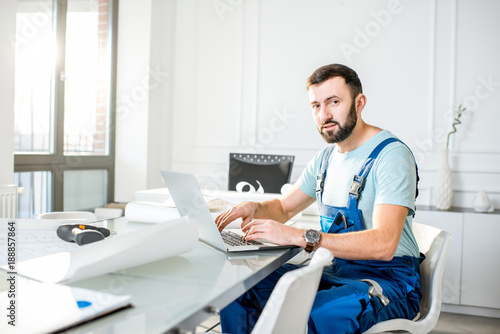 Handsome repairman or foreman in uniform working with laptop and architectural drawings at the office