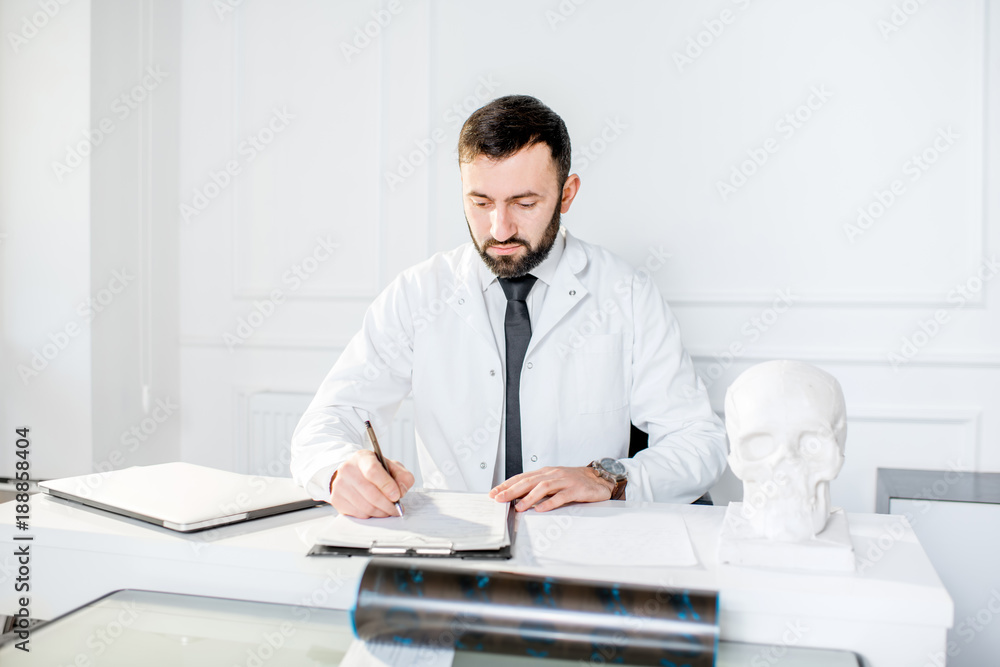 Handsome male doctor in the medical gown writing scientific works sitting in the beautiful white office interior