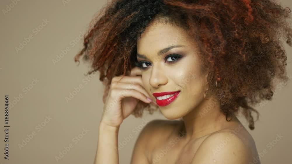 Portrait of cheerful ethnic woman with red lips touching curly hair and looking at camera in studio. Portrait of cheerful ethnic woman with red lips touching curly hair and looking at camera in studio.