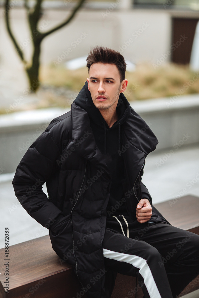 Modern outfit. Fashionable man sit on the bench and look around. Autumn and winter clothing style. Jacket with white sneakers. Modern outfit. Fashionable man sit on the bench and look around. Autumn and winter clothing style. Jacket with white sneakers.
