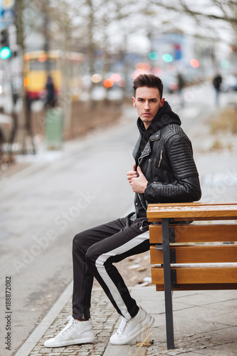 Modern outfit. Fashionable man sit on the bench and look around. Autumn and winter clothing style. Jacket with white sneakers.