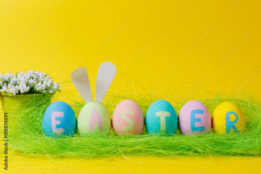 Row of six colorful pastel monophonic painted Easter eggs with inscription Easter, fun bunny ears, white flowers in bucket in green grass isolated on yellow background. Copy space for advertisement.