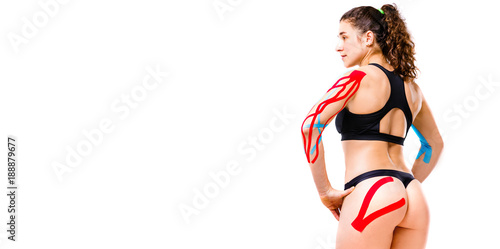 sexy female athlete with beautiful booty stands half sideways on white background in black top and bikini.On body is pasted kinesiology tape.Colored tapes for treatment of muscles,tendons in athletes