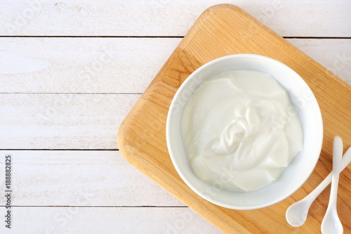 Greek yogurt in a white bowl, above view on a natural wood cutting board against white wood.