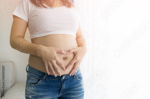 Pregnant Asian Woman holding her hands in a heart shape on her belly near window at home.copy space.