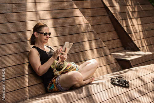 Woman sitting at wood bench in shade, using smartphone, touching touchscreen;