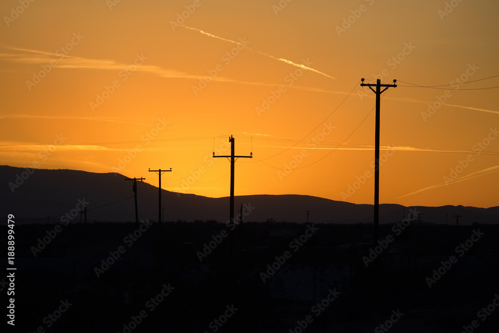 A row of telephone poles provide contrast in front of an orange sunset over a mountain range.