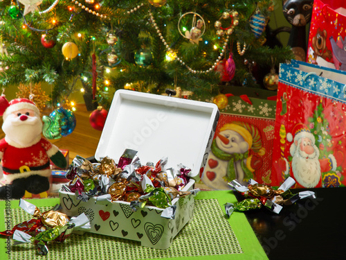 Decoupage box full of traditional Hungarian parlour candies. Beautifully decorated Christmas tree, gift bags under it, and a Santa Claus puppet are in a blurry background. Selective focus.
