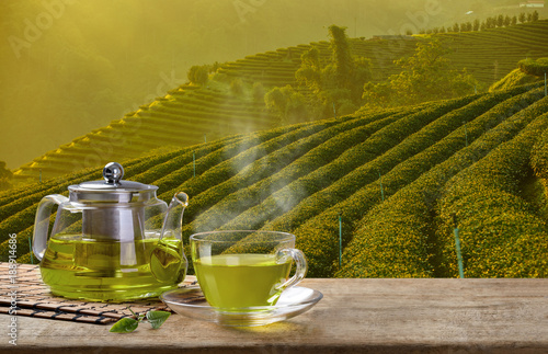 Warm cup of green tea and glass jugs or jars and green tea leaf on wooden table with the tea plantations background