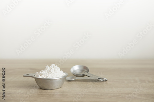 MEASURING CUP WITH FLOUR AND WRITING SPACE White flour in a measuring cup, and measuring spoon are on a wood table. There is writing space in the picture.